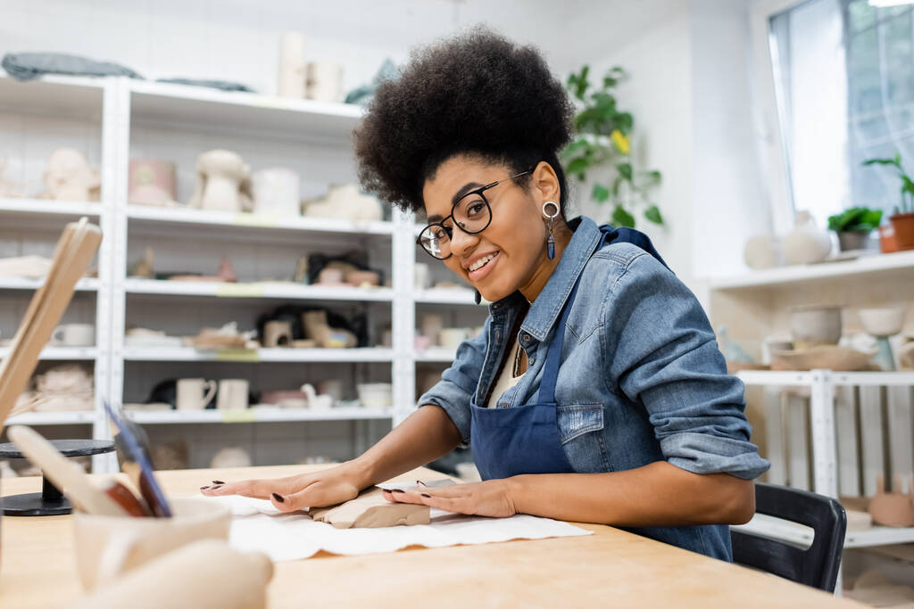 veselá africká americká žena v zástěře a brýlích tvarování hliněný kus s rukama během keramiky třídy - Fotografie, Obrázek
