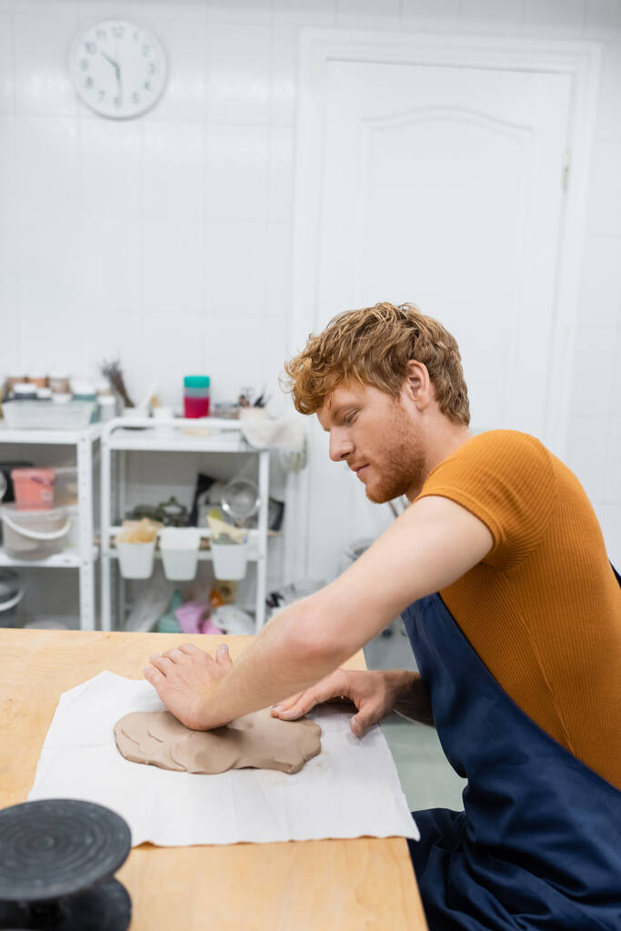 side view of redhead man in apron shaping clay piece with hand in pottery workshop - Photo, Image