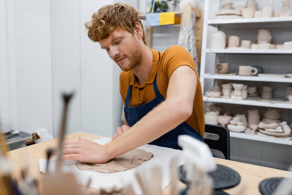 vousatý ryšavý muž v zástěře tvarování hliněný kus s rukou v keramické dílně - Fotografie, Obrázek