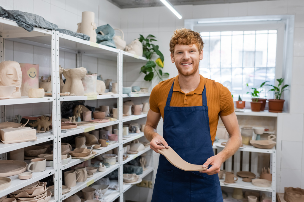positive redhead man in apron holding rectangle shape clay piece in hands near sculptures on rack - Photo, Image