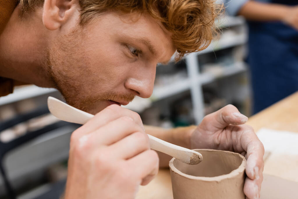 concentrated man with red hair holding shaper while modeling clay cup in pottery workshop - Photo, Image