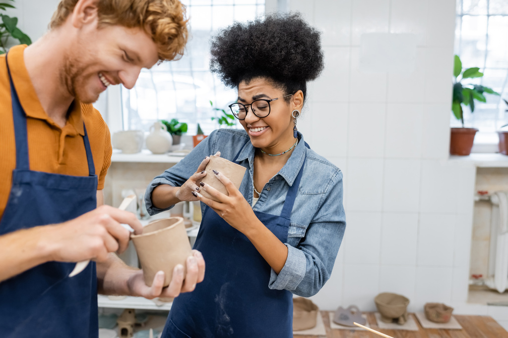 cheerful african american woman in eyeglasses molding clay cup with happy redhead boyfriend in pottery studio  - Photo, Image