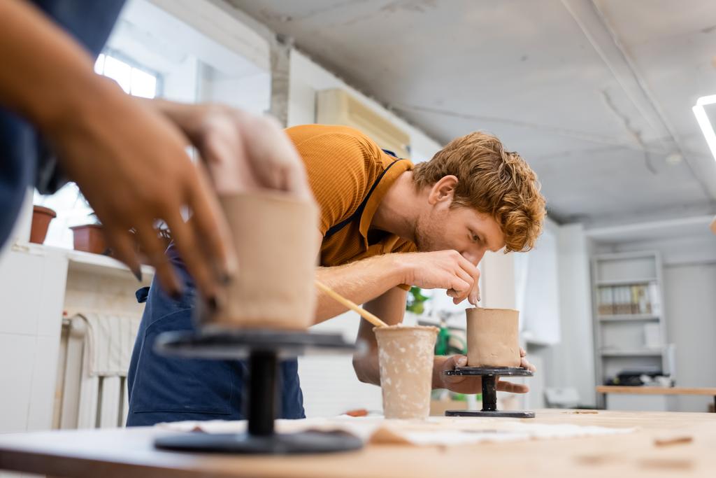 Redhead man forming clay sculpture near blurred african american girlfriend in pottery studio  - Photo, Image