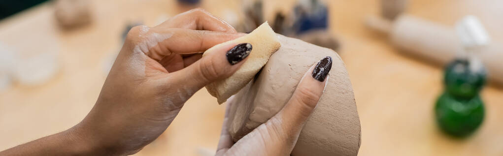 Vista recortada de artesano afroamericano sosteniendo esponja húmeda y arcilla en taller de cerámica, pancarta  - Foto, Imagen