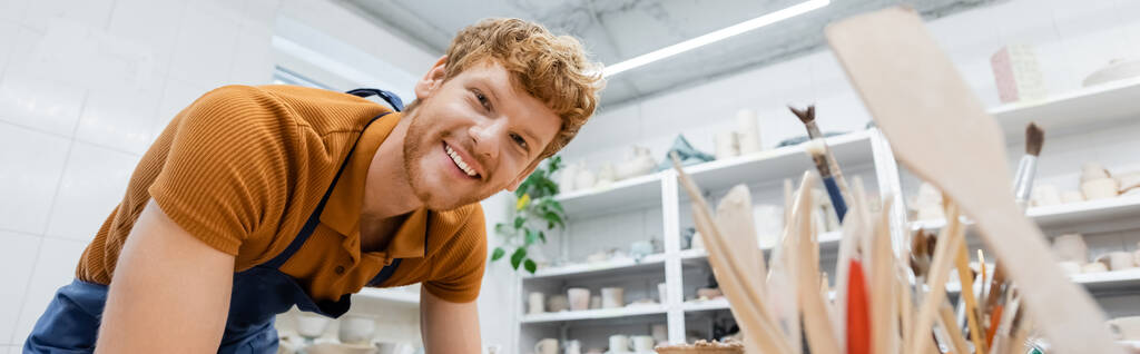 Artesano alegre mirando la cámara cerca de herramientas de cerámica borrosa en el taller, pancarta  - Foto, imagen