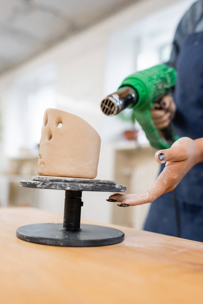Vista recortada de artesano afroamericano sosteniendo pistola de calor borrosa cerca de escultura de arcilla en taller de cerámica  - Foto, Imagen
