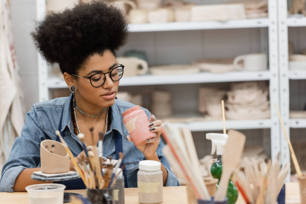 Artisanat afro-américain en lunettes tenant un bocal avec de la peinture près de pinceaux flous et sculpture en argile dans un atelier de poterie  - Photo, image