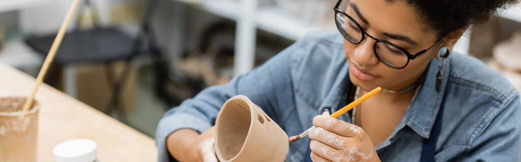 Молода афроамериканська ремісниця в окулярах малює глиняний виріб в керамічній майстерні, банер.  - Фото, зображення