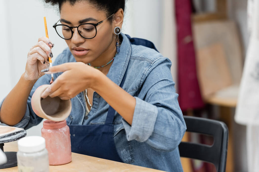 Artesana afroamericana en anteojos pintando sobre producto cerámico cerca de frascos con pintura en estudio de cerámica  - Foto, Imagen