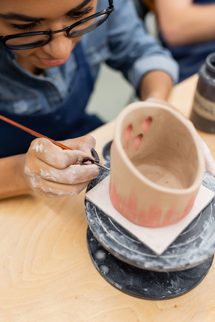 Giovane donna afroamericana in occhiali da vista pittura su prodotto argilla sfocata in studio di ceramica  - Foto, immagini