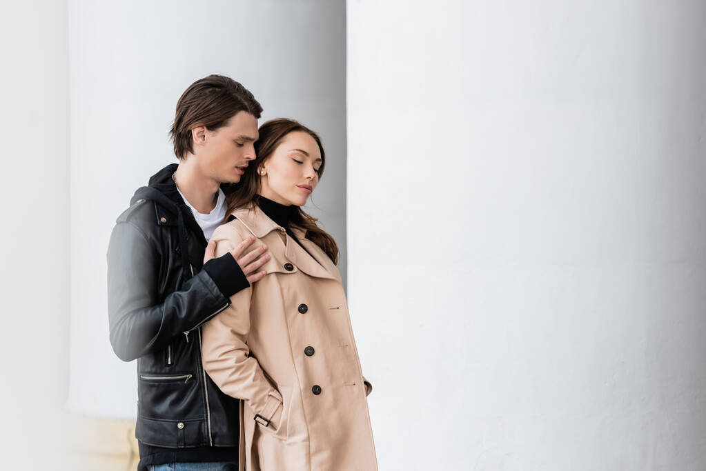 stijlvolle man in jas knuffelen schouders van sensuele vriendin staan met de handen in zakken van trench jas in de buurt kolommen - Foto, afbeelding