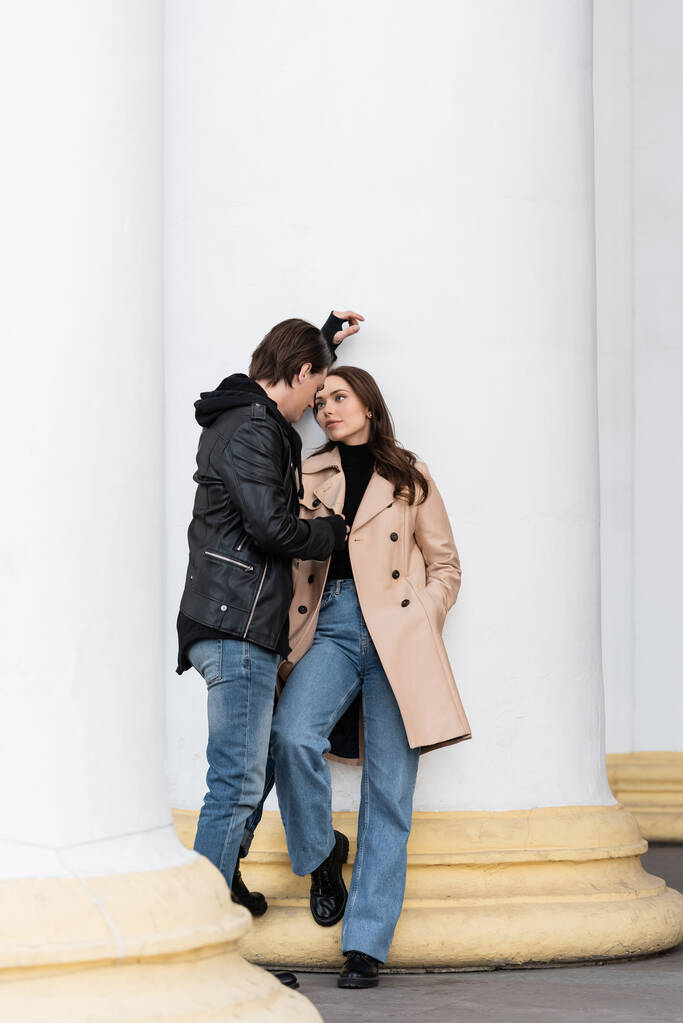 full length of young man in stylish jacket flirting with pretty woman in beige trench coat standing with hand in pocket - Photo, Image