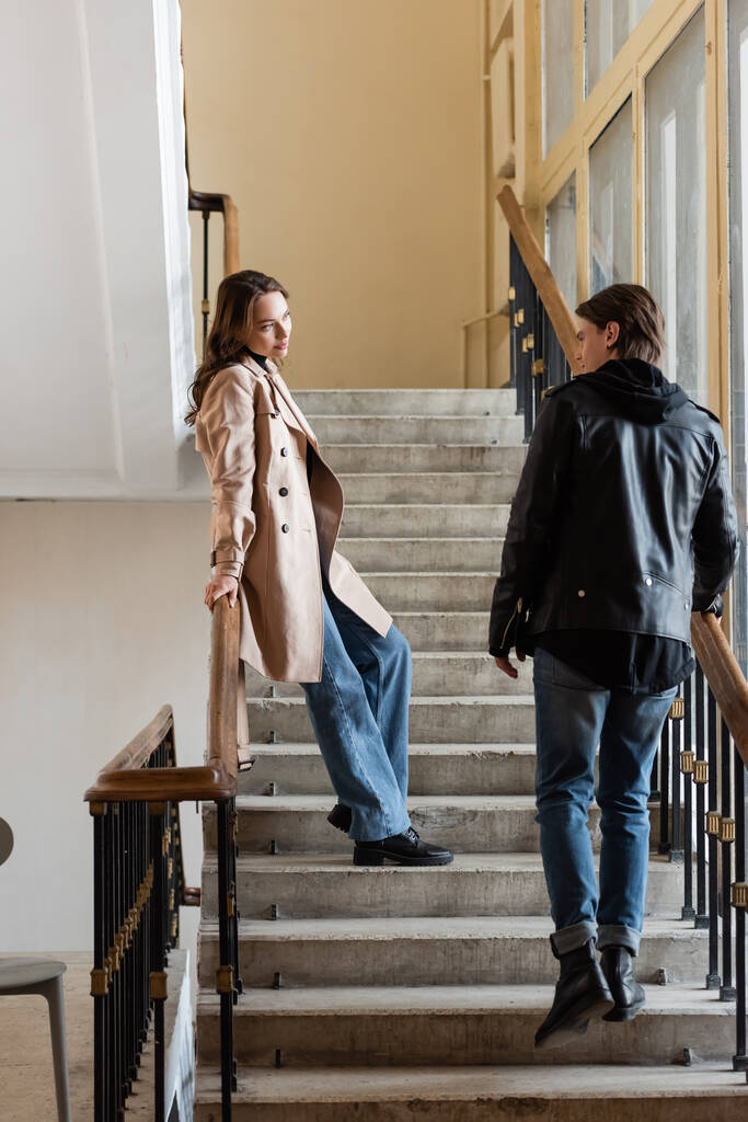 full length of young man in stylish outfit looking at pretty girlfriend in beige trench coat standing on stairs  - Photo, Image