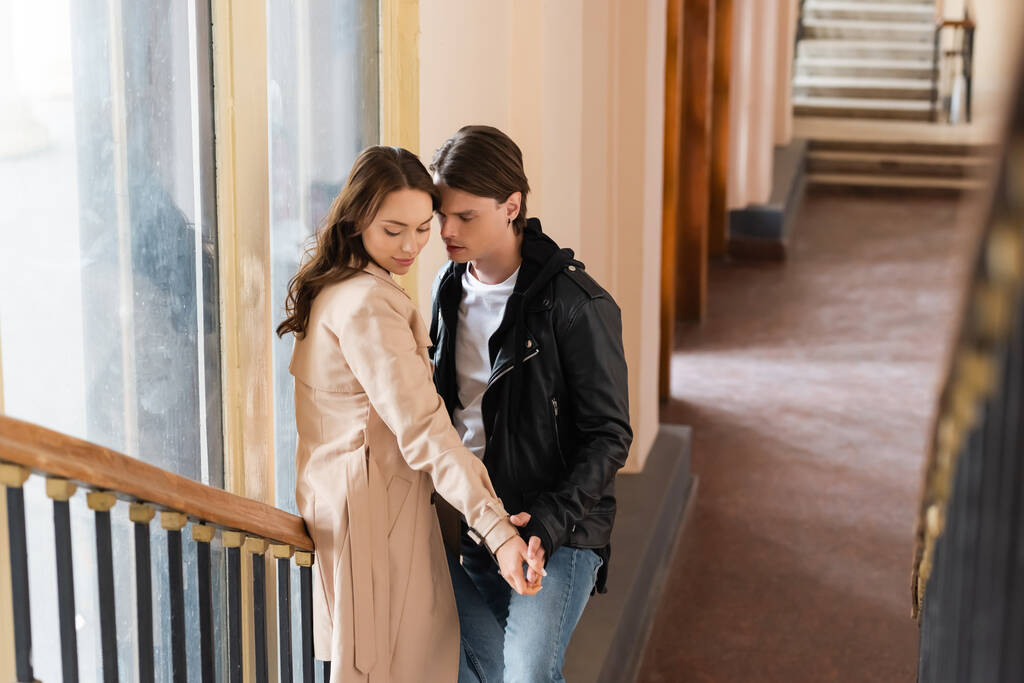 fiatal férfi fekete dzsekiben flörtöl barátnőjével őszi kabátban, miközben fogja a kezét a lépcsőn  - Fotó, kép