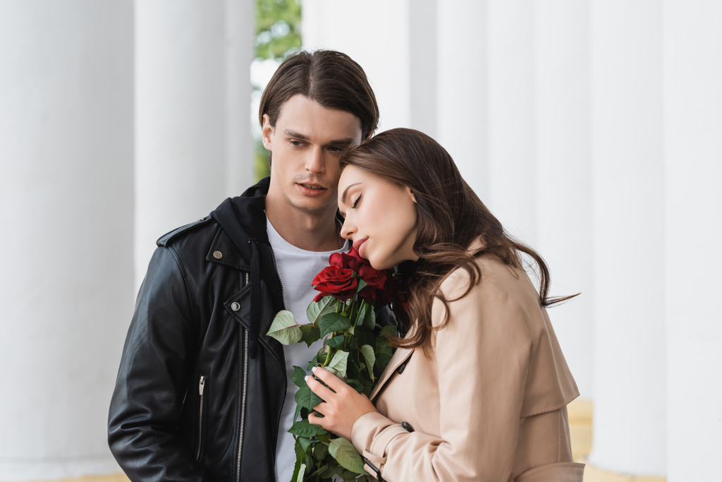bonita jovem mulher cheirando rosas vermelhas perto de namorado elegante na jaqueta - Foto, Imagem