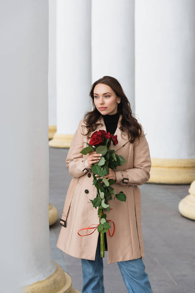 hübsche junge Frau im beigen Trenchcoat, rote Rosen in der Hand und wegschauend - Foto, Bild