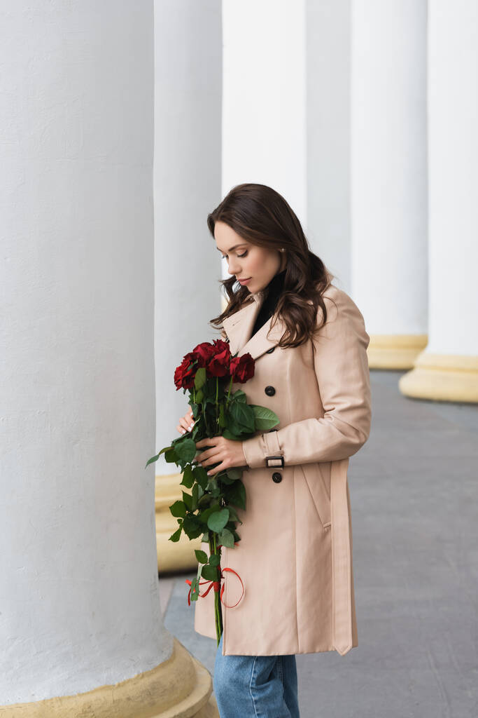 bonita mujer joven en gabardina beige mirando rosas rojas florecientes - Foto, imagen