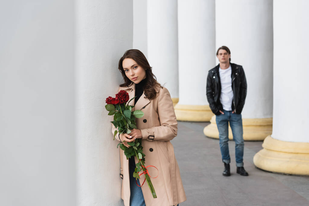 гарна жінка в бежевому траншеї пальто тримає червоні троянди біля хлопця на розмитому фоні
  - Фото, зображення