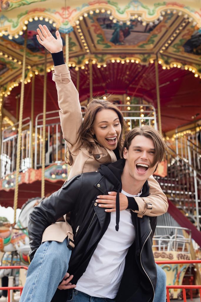 excited man in black jacket piggybacking happy girlfriend in amusement park - Photo, Image