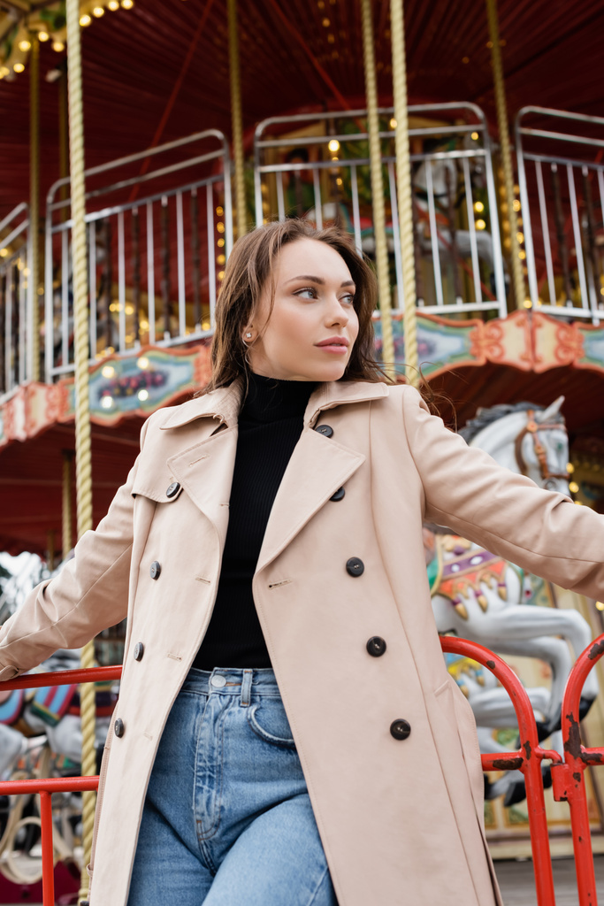 brunette nuori nainen beige trenssi takki ja farkut seisoo lähellä karuselli huvipuistossa - Valokuva, kuva