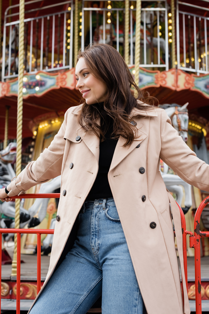 cheerful young woman in beige trench coat and jeans standing near carousel in amusement park - Photo, Image