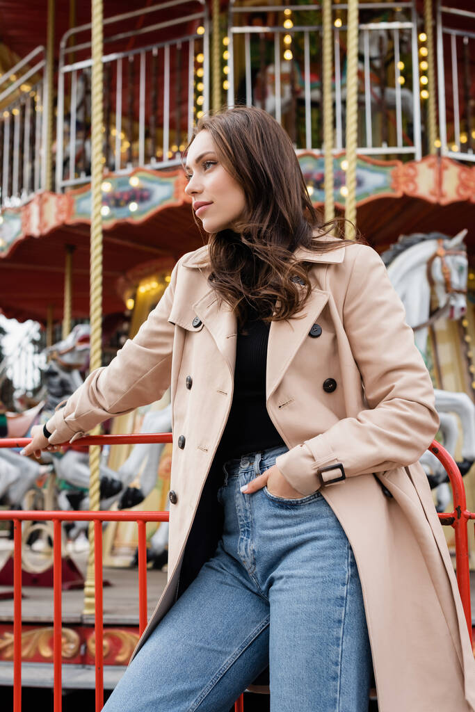 Jeune femme en trench coat beige et jeans debout la main dans la poche près du carrousel dans le parc d'attractions - Photo, image