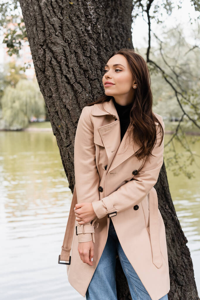 pleased young woman in beige trench coat leaning on tree trunk in park - Photo, Image