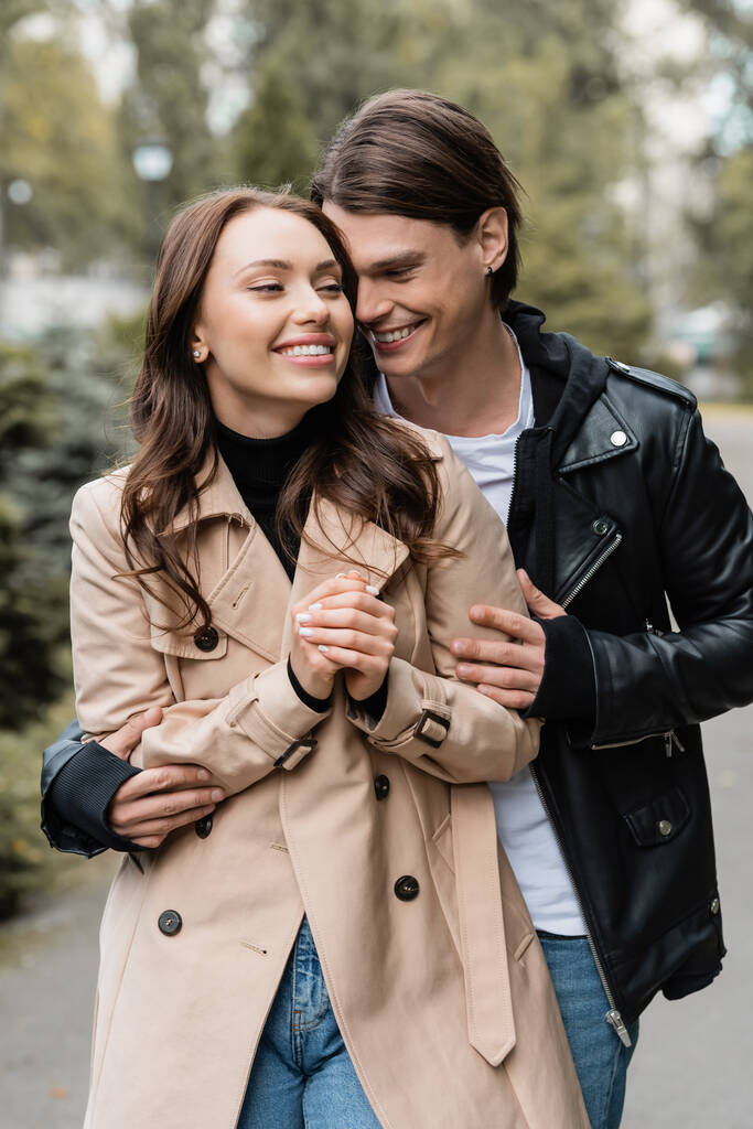 радісний молодий чоловік обіймає щасливу дівчину в траншеї пальто на відкритому повітрі
 - Фото, зображення