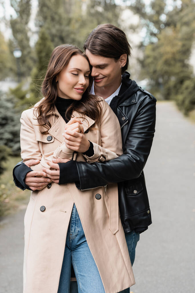šťastný mladý muž objímání potěšené přítelkyně v trench kabát venku - Fotografie, Obrázek