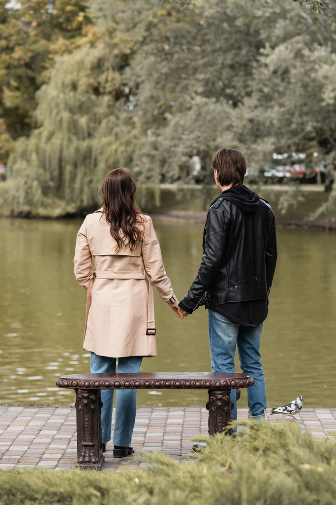 πίσω όψη του νεαρού ζευγαριού σε φθινοπωρινά ρούχα κρατώντας τα χέρια, ενώ στέκεται κοντά στη λίμνη στο πάρκο - Φωτογραφία, εικόνα