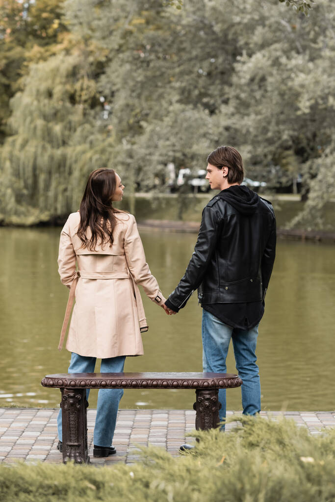 vue arrière du jeune couple dans des tenues élégantes tenant la main tout en se tenant près du lac dans le parc automnal - Photo, image