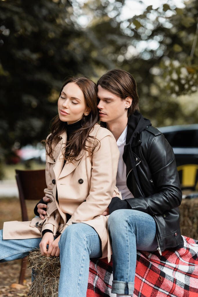stijlvolle man in zwart jasje knuffelen jonge vriendin met gesloten ogen zitten in park - Foto, afbeelding