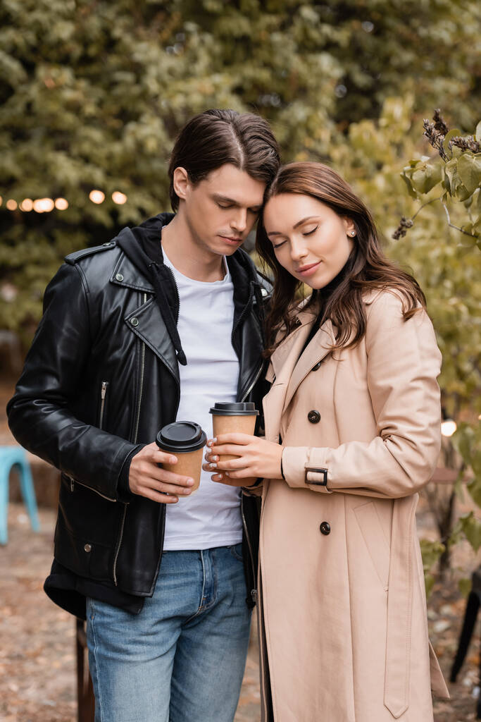 jonge vrouw en man in stijlvolle outfits staan met gesloten ogen en houden koffie te gaan  - Foto, afbeelding