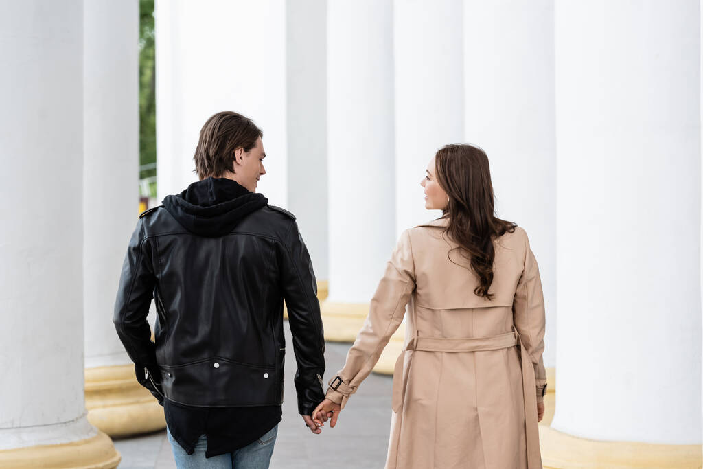 feliz joven hombre y mujer tomados de la mano mientras camina cerca de columnas blancas - Foto, Imagen