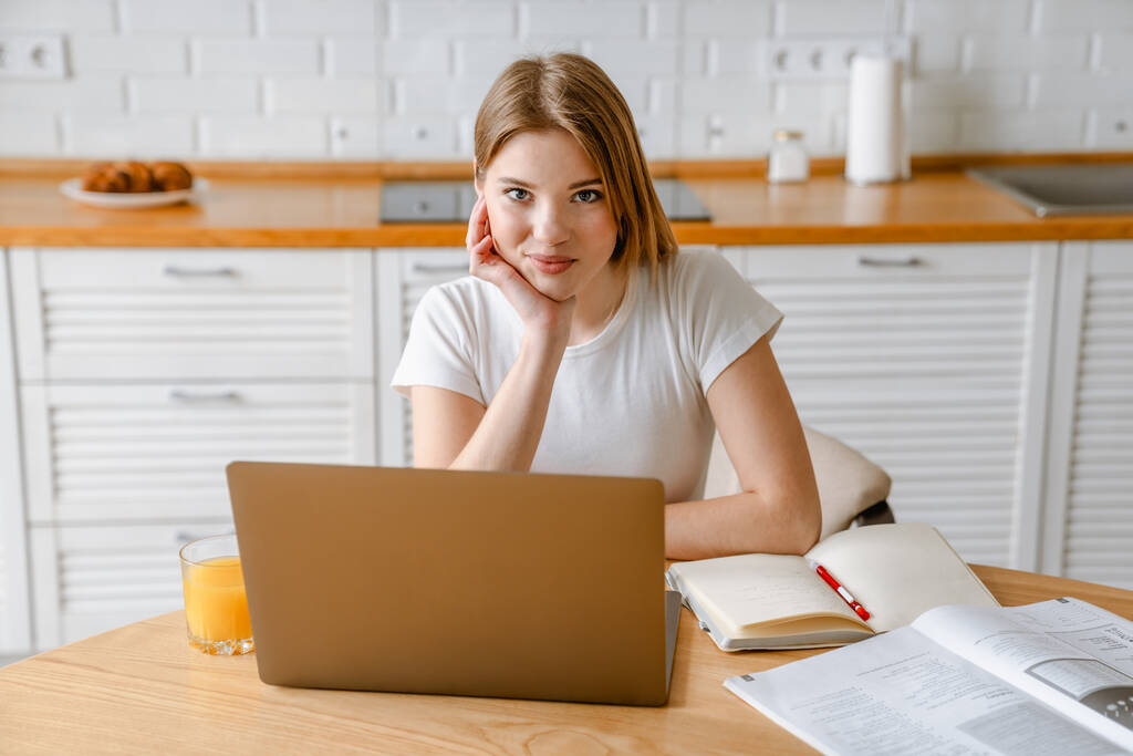 Sorridente giovane donna bianca bionda seduta con computer portatile al tavolo della cucina - Foto, immagini