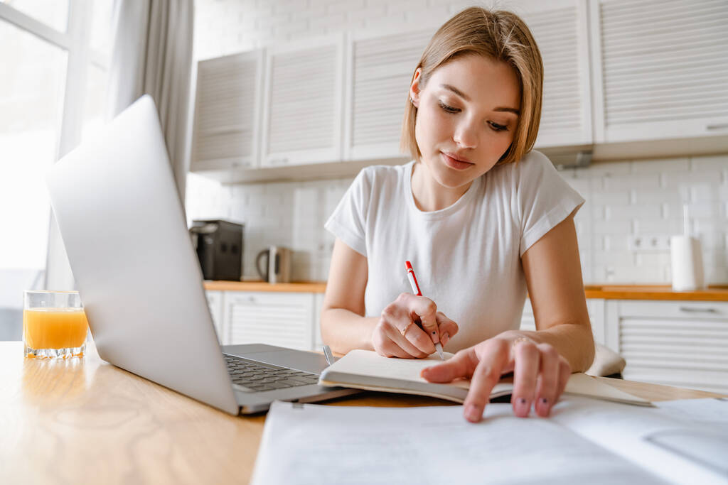 Улыбающаяся молодая женщина, занимающаяся с ноутбуком, сидит дома, делает заметки - Фото, изображение