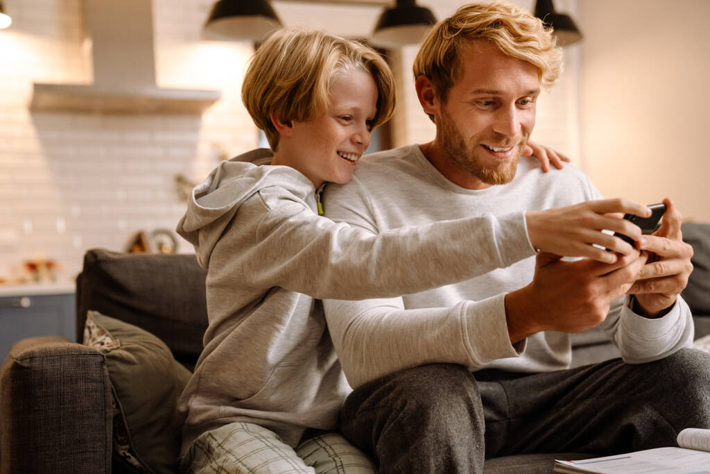Ginger férfi mobiltelefont használ, miközben otthon viccelődik a fiával. - Fotó, kép