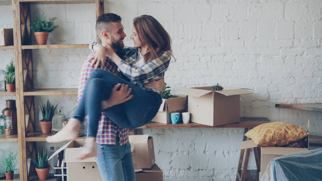 Glücklicher starker Mann wirbelt seine aufgeregte Frau auf und küsst sie beim Umzug in ein neues Haus nach dem Kauf. Romantische Beziehung, Menschen und Wohnkonzept. - Foto, Bild