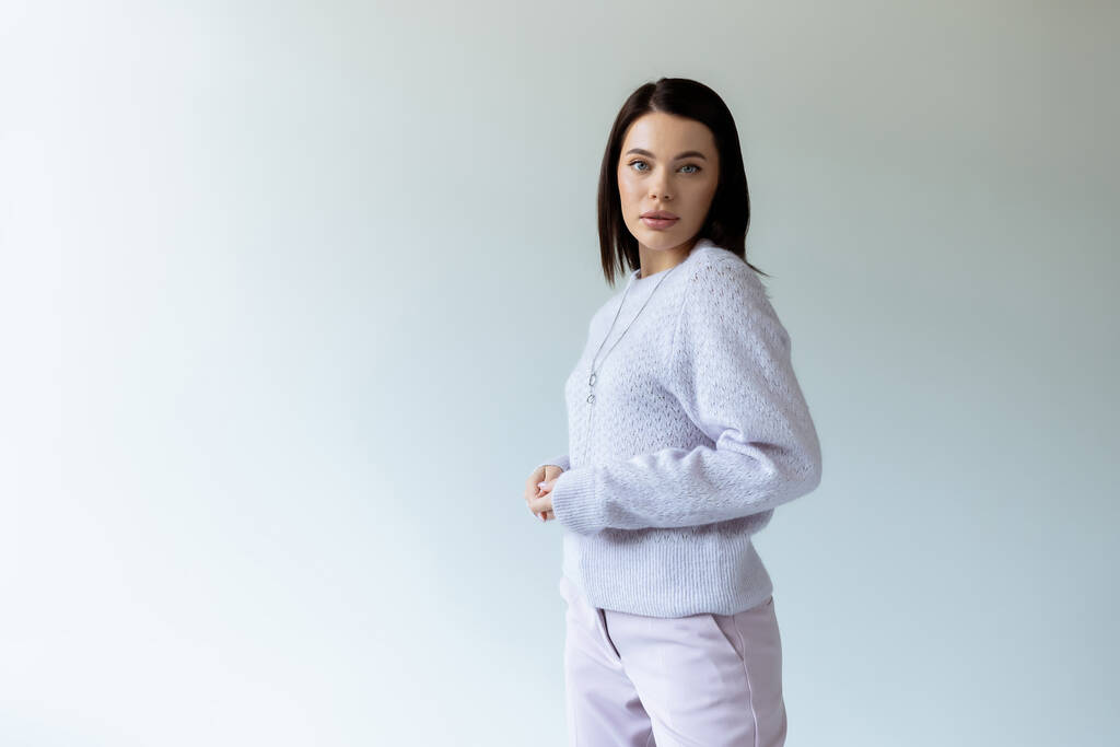 brunette woman in knitted sweater and trousers looking at camera on grey background - Photo, Image