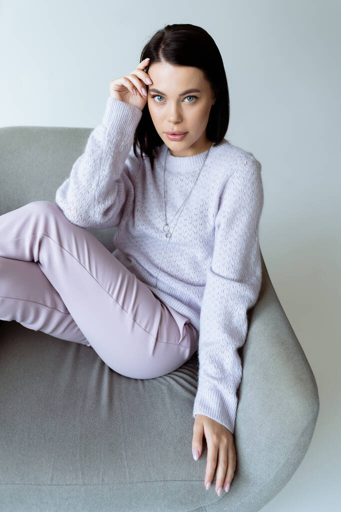 brunette woman in warm sweater and trousers sitting on comfortable armchair  isolated on grey - Photo, Image