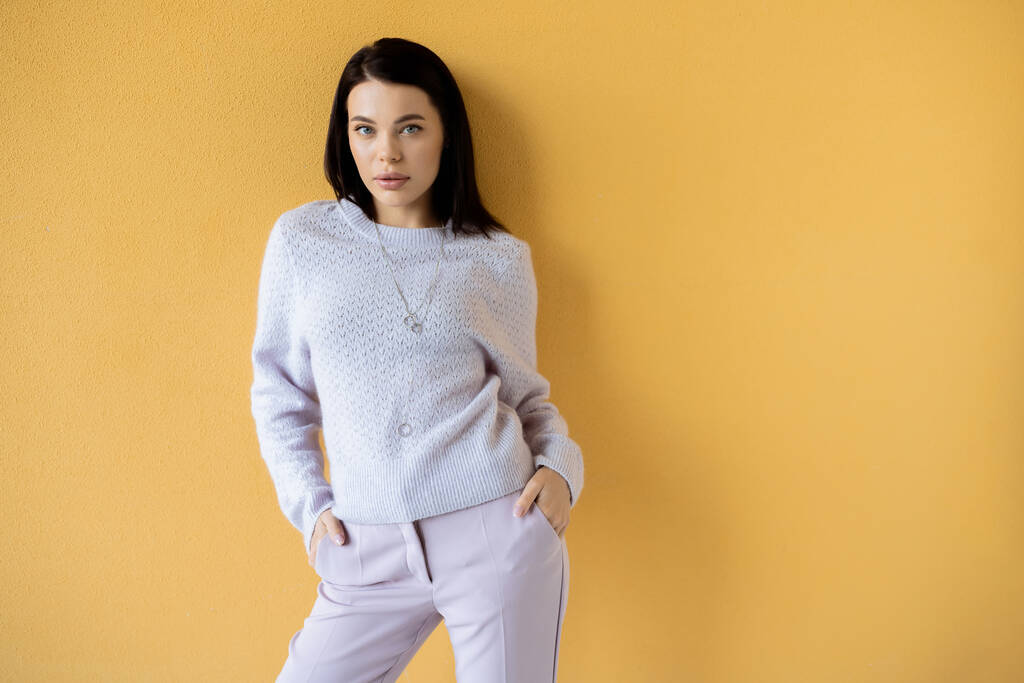 brunette woman in soft knitted sweater standing with hands in pockets of trousers on yellow background - Photo, Image