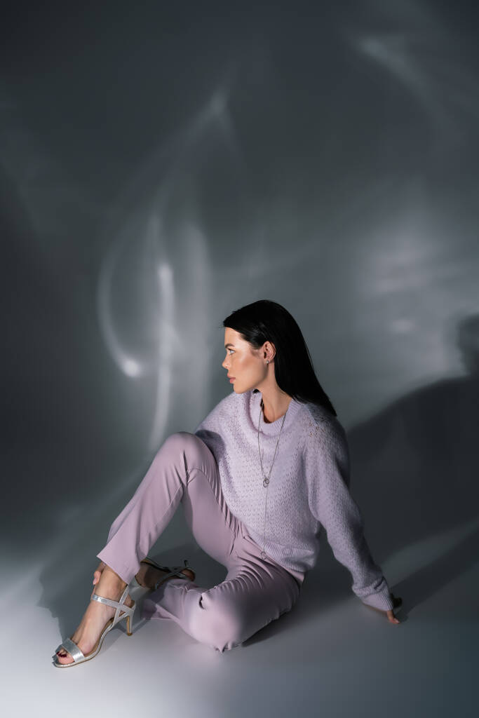 πλήρες μήκος της μελαχρινής γυναίκας σε άνετο πουλόβερ και σανδάλια κάθεται σε γκρι φόντο με φωτισμό  - Φωτογραφία, εικόνα