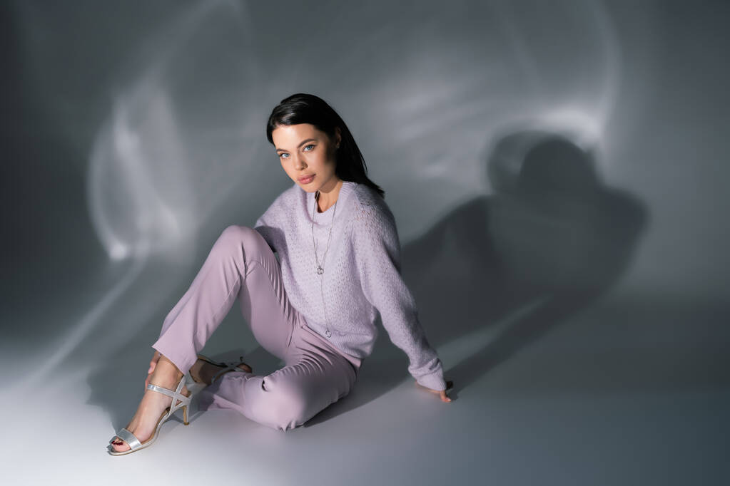 joven morena en ropa de moda y sandalias sentado sobre fondo gris con iluminación - Foto, Imagen