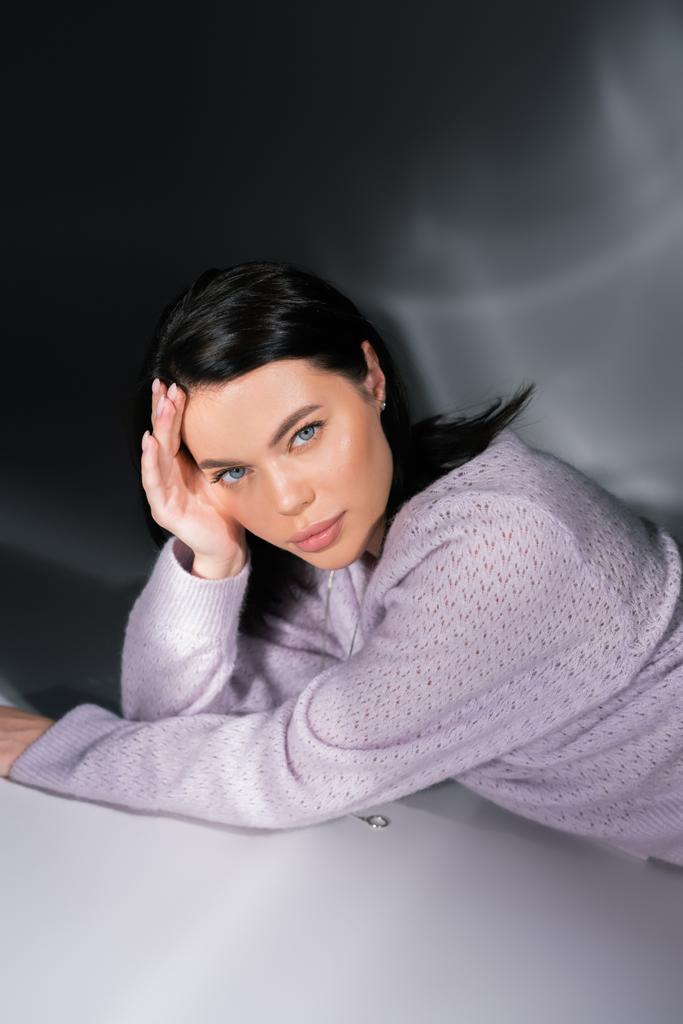 брюнетка женщина в трикотажном свитере лежит на сером фоне с освещением - Фото, изображение