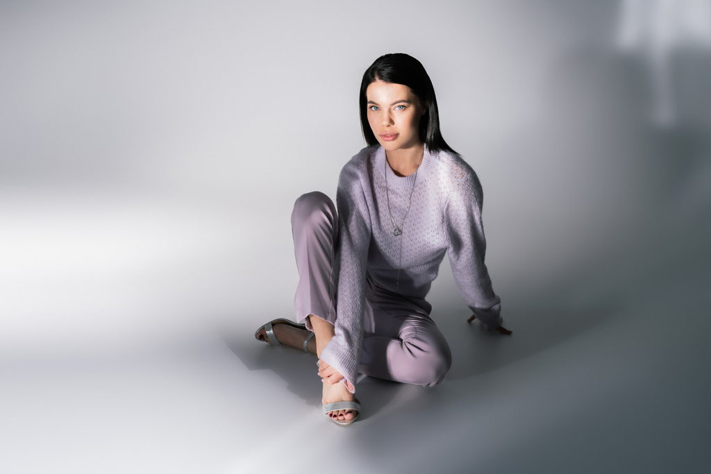 full length of young and stylish woman in sweater and trousers sitting on grey background - Photo, Image