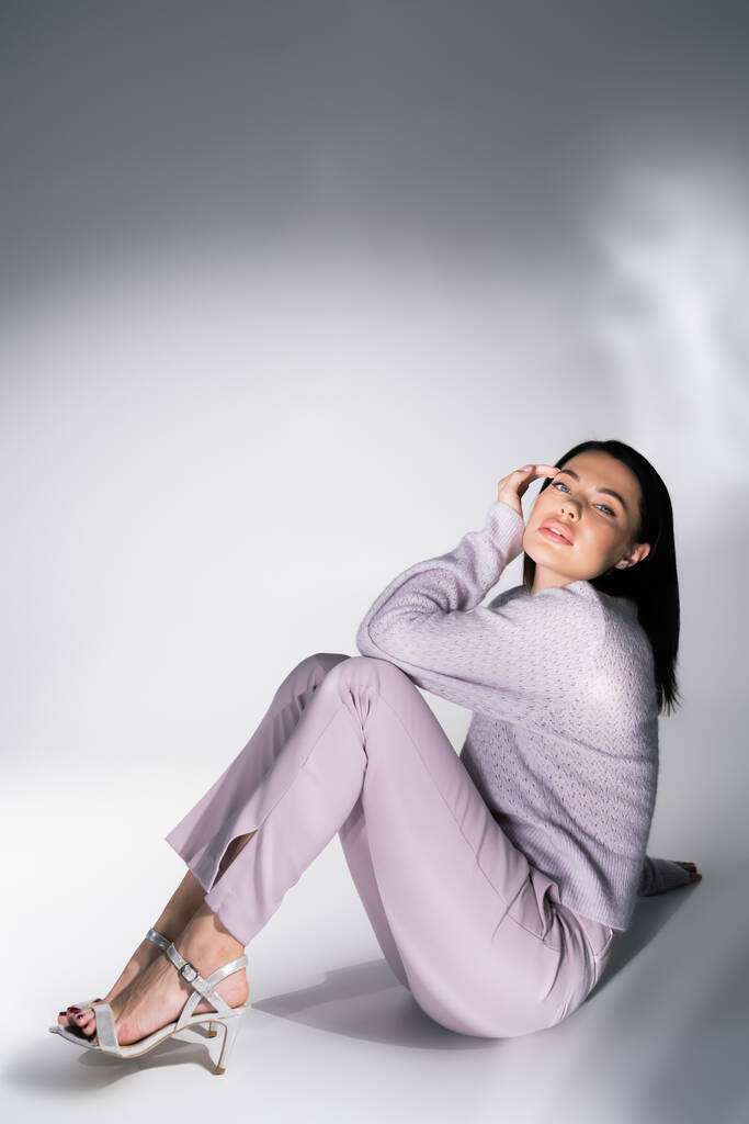 mujer encantadora en suéter de punto y pantalones sentados y mirando a la cámara sobre fondo blanco y gris - Foto, Imagen