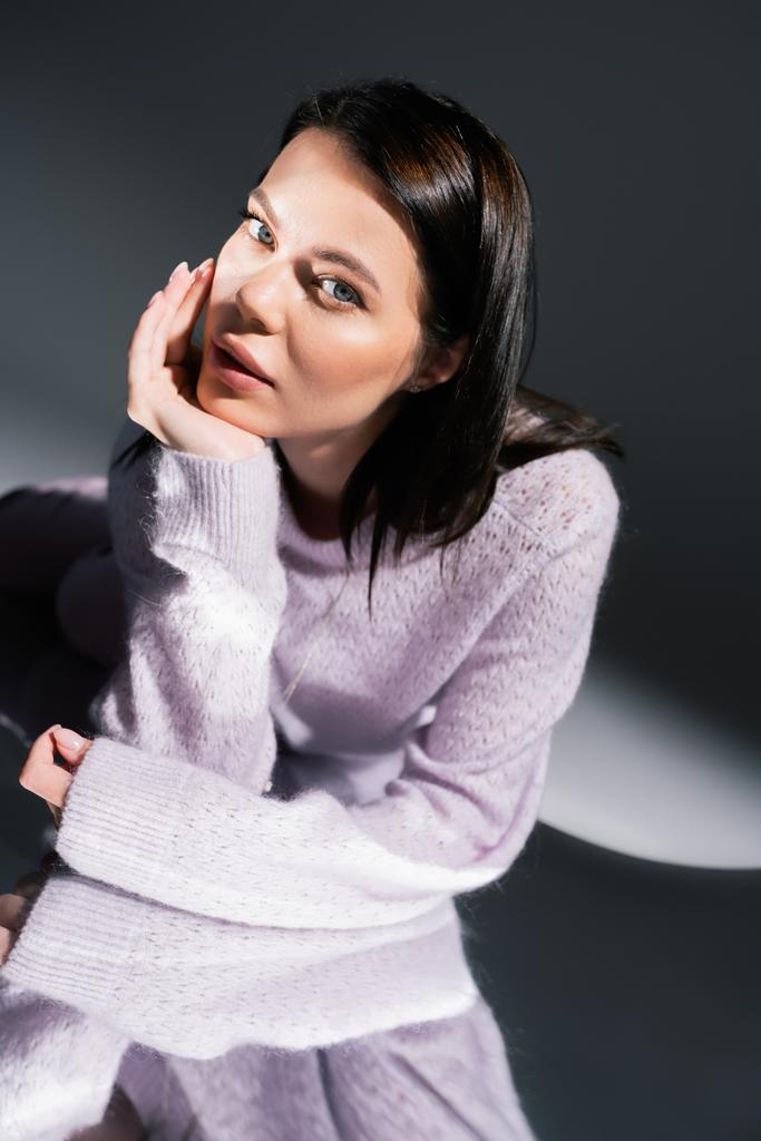 αισθησιακή μελαχρινή γυναίκα σε ζεστό πουλόβερ κοιτάζοντας κάμερα, ενώ βρίσκεται σε γκρι φόντο με φωτισμό - Φωτογραφία, εικόνα