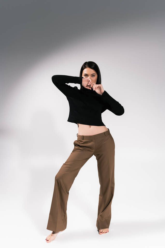 full length of barefoot woman in brown pants and black sweater posing with hands near face on grey background - Photo, Image
