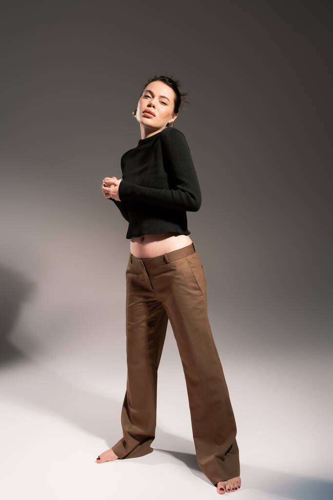 брюнетка босиком женщина в коричневых брюках и черный свитер стоя на сером фоне - Фото, изображение