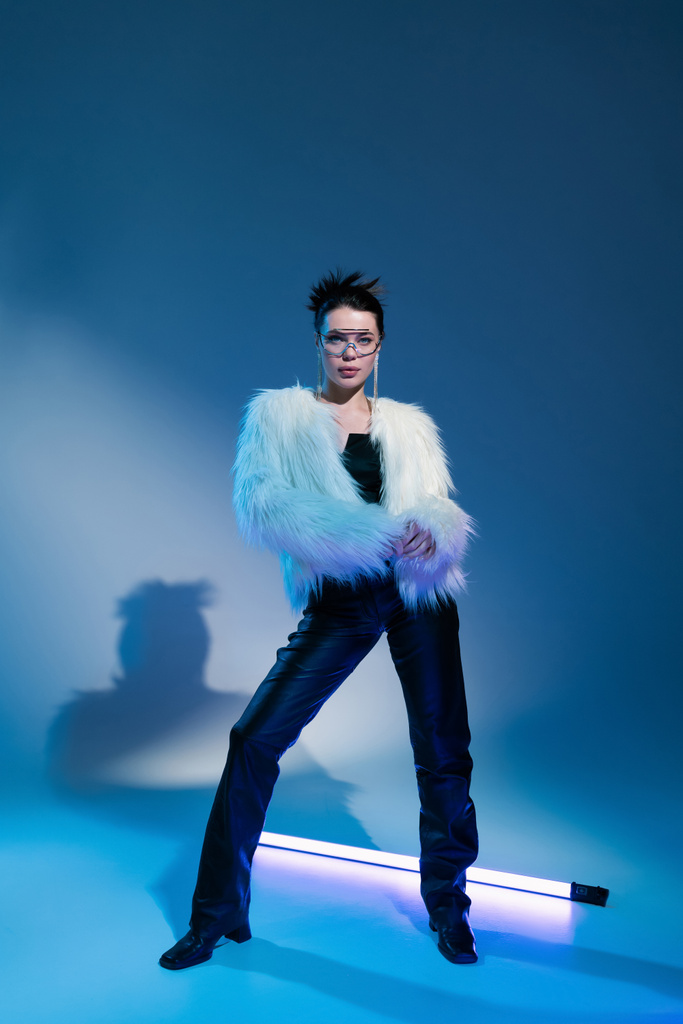 full length of fashionable woman in white faux fur jacket posing near fluorescent lamp on blue background - Photo, Image